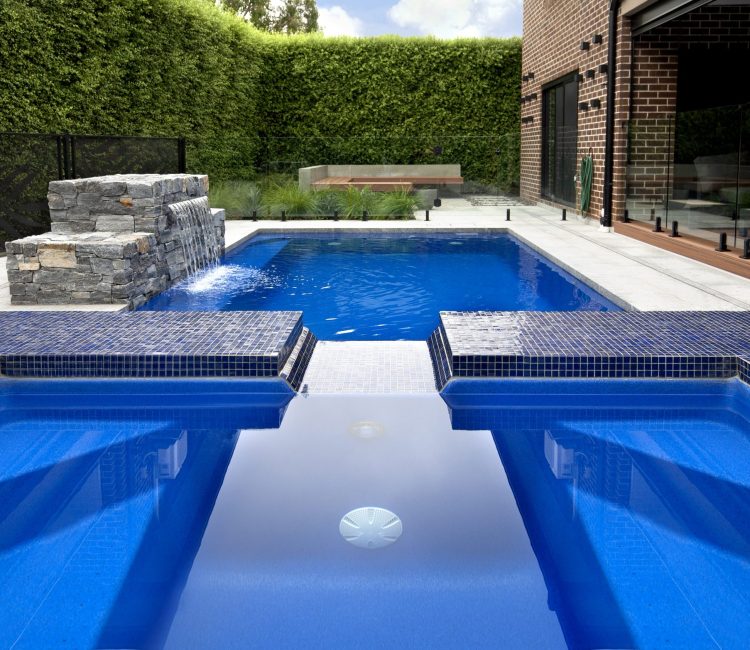 Fibreglass pool and spa in Berwick, Melbourne, featuring deep blue water, blue mosaic tile detailing, stacked stone water feature, frameless glass fencing, and established hedge backdrop with brick home.