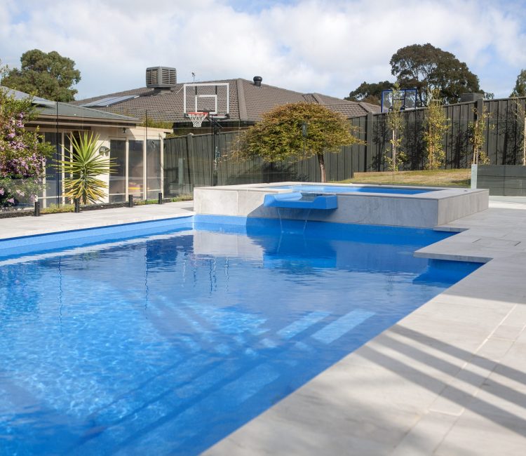 Fibreglass pool with elevated spa and water spillway in Botanic Ridge, Melbourne, featuring blue water, light grey paving, frameless glass fencing, flowering shrubs, and a basketball hoop in a spacious family backyard.