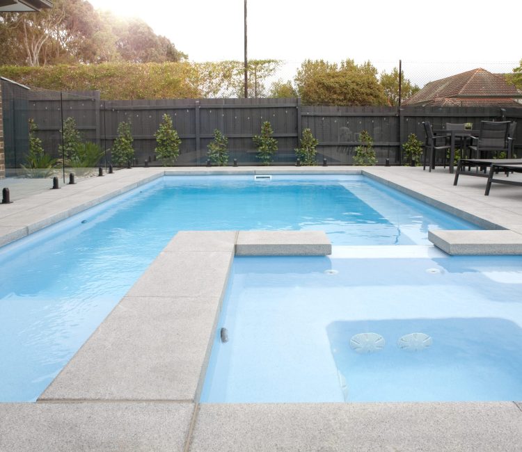 Fibreglass pool and spa combination in Lysterfield, Melbourne, with light blue water, grey paving, frameless glass fencing, sun lounges, and newly planted hedging along the fence line.